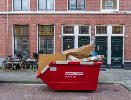 A large red skip bin positioned on the pavement in front of a row of brick terraced houses in Marylebone, London. The skip is filled with mixed waste materials including flattened cardboard boxes, plastic wrapping, and other household rubbish, with some items visibly protruding over the top edge. To the left of the skip, there are two bicycles leaning against the wall of the building, one with a black frame and the other with metallic components. The buildings behind have tall, narrow windows with metal security bars and white window frames, along with dark green front doors featuring mail slots and doorbells. The overall scene suggests a typical urban setting with ongoing rubbish clearance activity, aligning with professional waste removal services in the Marylebone area. The environment appears clean and orderly, with natural daylight illuminating the scene, emphasizing the textures of the brickwork and the materials inside the skip, reflecting a typical house clearance process carried out by Garden Clearance Marylebone.