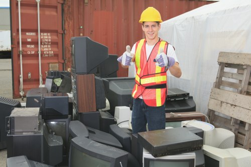A young male waste management worker wearing a yellow safety helmet, high-visibility vest with orange and yellow stripes, and purple work gloves standing outdoors in front of a collection of discarded electronic devices and office equipment. The electronic waste includes various old CRT monitors, computer towers, and printers, all stacked on a paved surface. Behind the worker, there is a large rust-colored shipping container and a wooden pallet leaning against a white wall or sheet. The scene appears to be part of a rubbish clearance operation, with the worker smiling and giving a thumbs-up gesture, indicating a successful rubbish removal process. The background suggests a practical, industrial environment typical of local rubbish collection or recycling sites in Marylebone, London, connecting the image to professional waste management services such as those offered by Garden Clearance Marylebone.