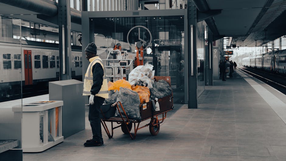 A pile of discarded household items placed on a paved driveway, consisting of broken wooden furniture, plastic containers, cardboard boxes, and miscellaneous waste materials. The wooden furniture appe