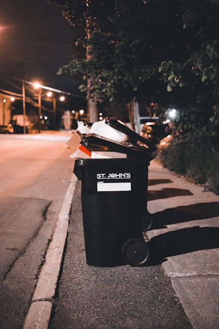 A collection of black rubbish bags and loose discarded items piled against a textured, off-white exterior wall beneath a small rectangular window set above a red metal door. The door features various