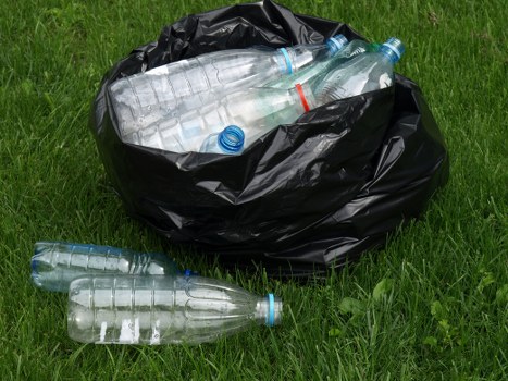 A black plastic rubbish bag placed on a grassy outdoor area contains several discarded clear plastic bottles, some with blue caps and one with a red band, partially visible inside the bag. Two additional empty plastic bottles lie on the grass in front of the bag, with their labels removed and caps still attached. The background features vibrant green grass, suggesting a garden or open space typical of residential areas in Marylebone. The image reflects the type of waste material that Garden Clearance Marylebone might handle for rubbish removal services, with a focus on plastic waste disposal. The scene is well-lit with natural daylight, emphasizing the textures of the plastic bottles and the grass. This visual highlights the importance of proper waste collection and recycling efforts in urban environments, supporting professional garden clearance and rubbish removal operations in central London neighborhoods such as Marylebone.
